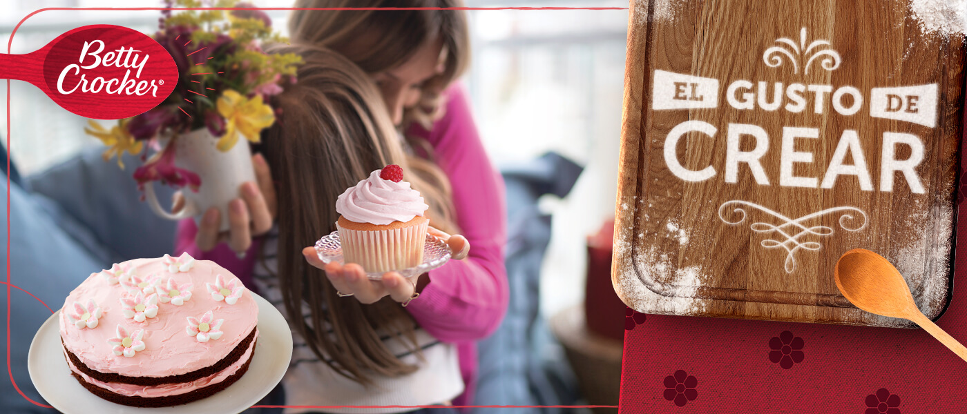 Adult woman hugging young child. White cupcake with red frosting on a plate. Chocolate cake with red frosting and white flowers on top on a plate. Wood cutting board and wooden spoon on a red table cloth. Purple and yellow flowers in vase.