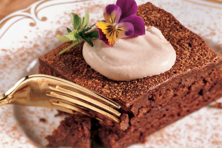 Chocolate mousse brownie with whipped cream on a white and gold plate. Gold colored fork and a purple/green flower on top of the whipped cream.