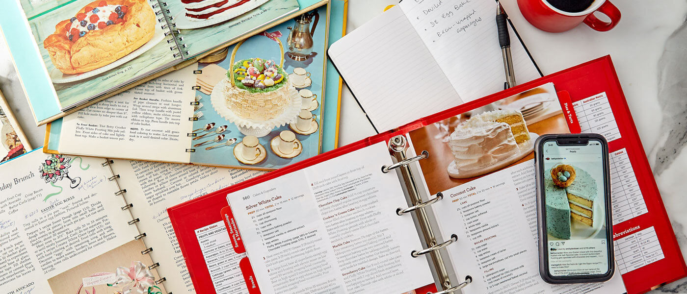 Lifestyle image of multiple Betty Crocker Cookbooks, a white notepad, cup of coffee and a cell phone displaying a image from social media